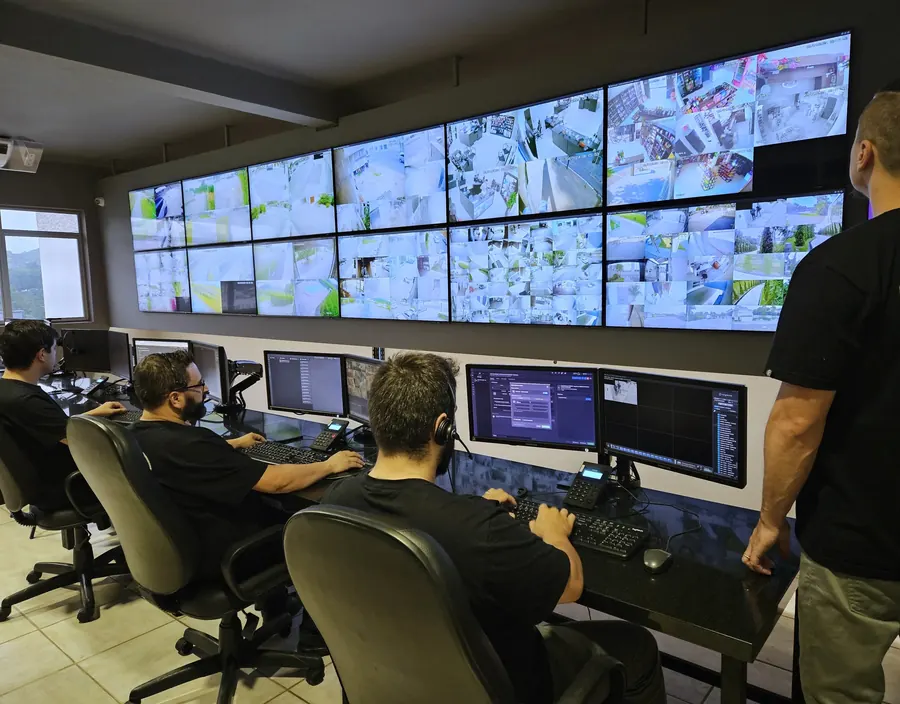Operadores da central de monitoramento trabalhando diante do videowall, com o supervisor acompanhando